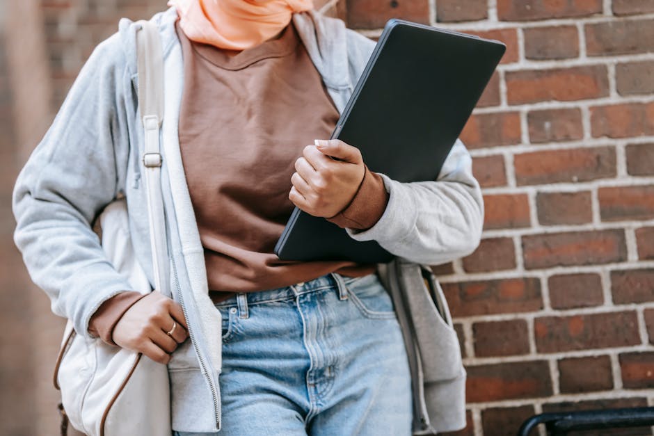 Unrecognizable ethnic woman in casual wear with bag and laptop standing beside brick wall on street