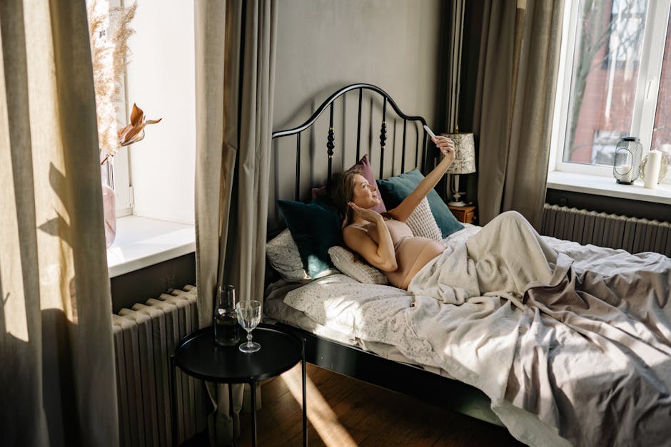 Pregnant woman lying on bed taking selfie, enjoying a peaceful morning in a sunlit bedroom.