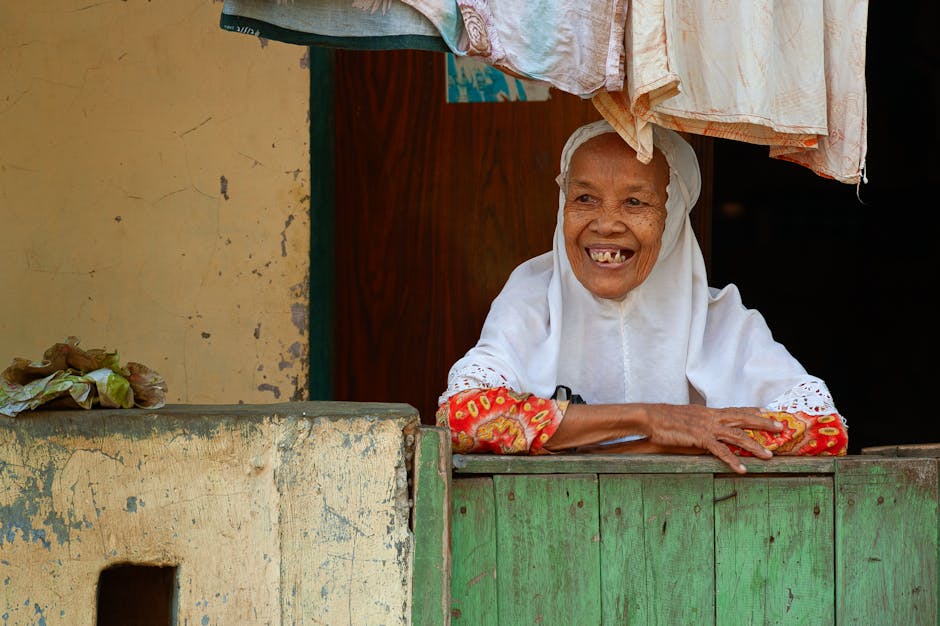 Joyful elderly woman in traditional attire smiling at a rustic window, exuding warmth.