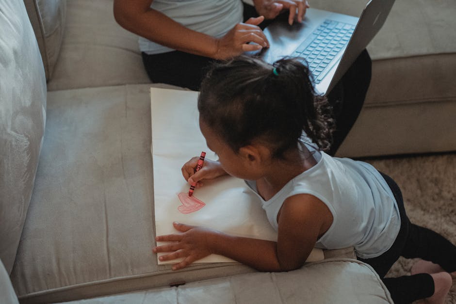 From above of ethnic little girl drawing on paper with wax crayon while mother using laptop sitting on sofa