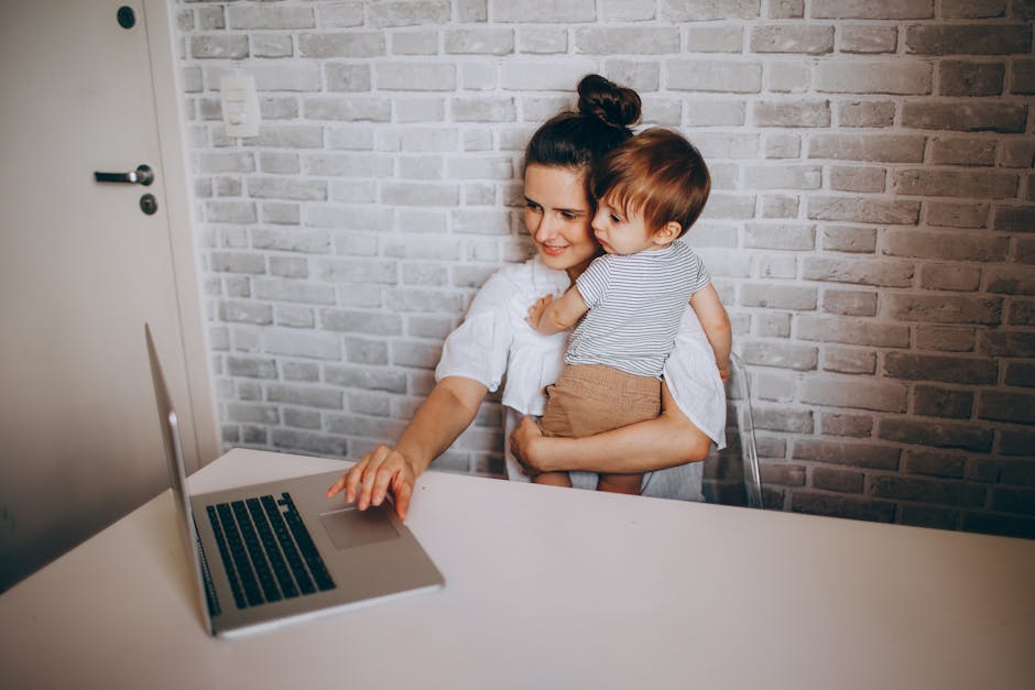 From above of smiling woman looking at laptop while sitting at table with little son on hands and working on remote project
