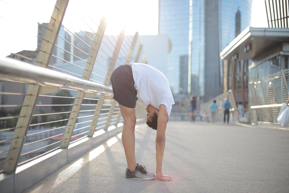 Side view of young African American man in earphones and sportswear listening to music and stretching body while performing standing forward bend exercise on street against blurred high rise buildings in downtown