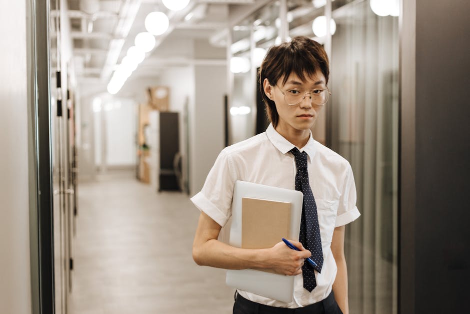 An adult in business attire with glasses walking through an office hallway holding a laptop and notebook.