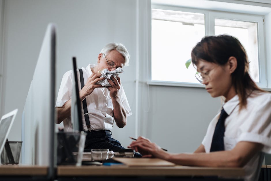 Two office employees on break. Senior man eating while another uses the computer.