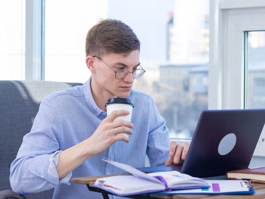 Adult wearing eyeglasses works remotely on a laptop while enjoying coffee indoors.