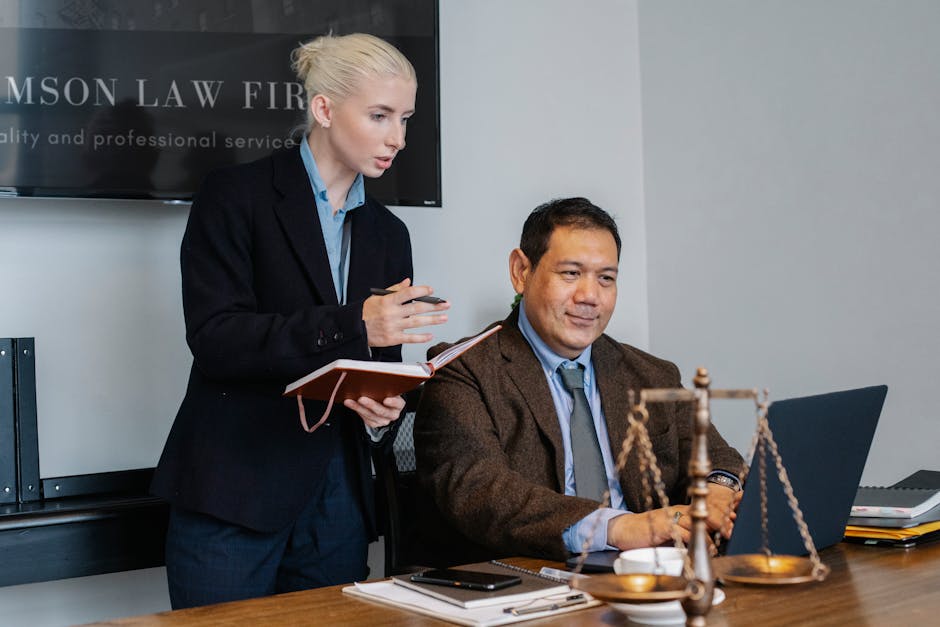Concentrated young female assistant taking notes in planner while standing at table near middle aged ethnic male colleague typing on laptop during work in law firm