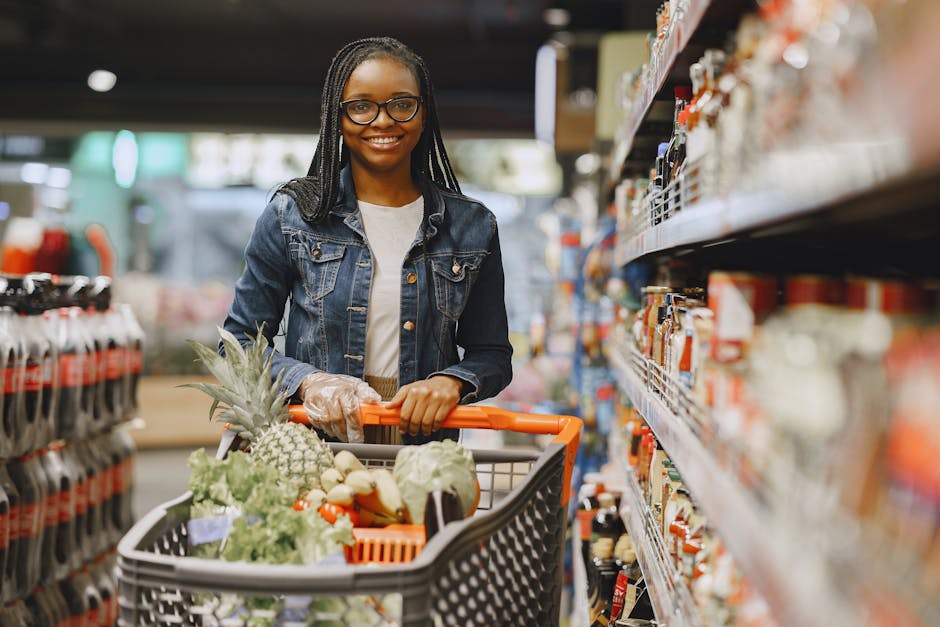 African American woman with glasses shopping in a grocery store aisle with a full cart of fresh produce.