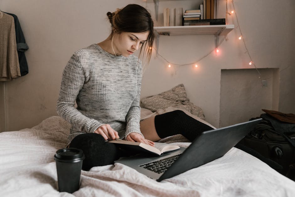 Young woman reading and studying on bed with a laptop in a cozy bedroom setting.