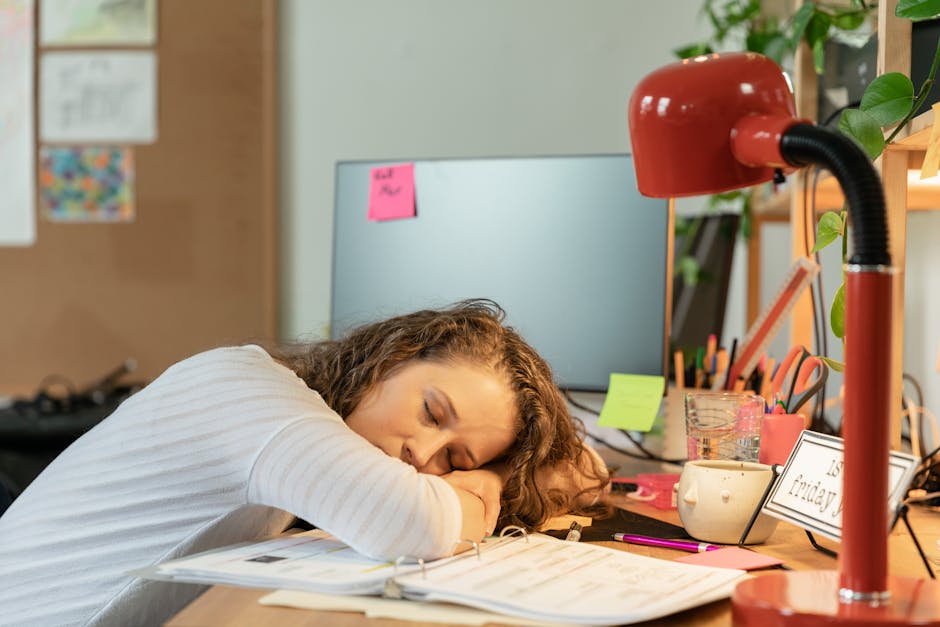 A woman sleeping at her office desk, surrounded by work documents and a red desk lamp.