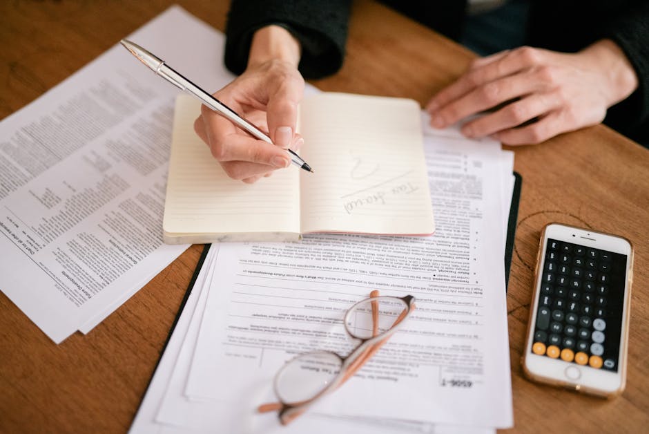 Close-up of a person writing in a notebook with documents and a calculator, managing finances.