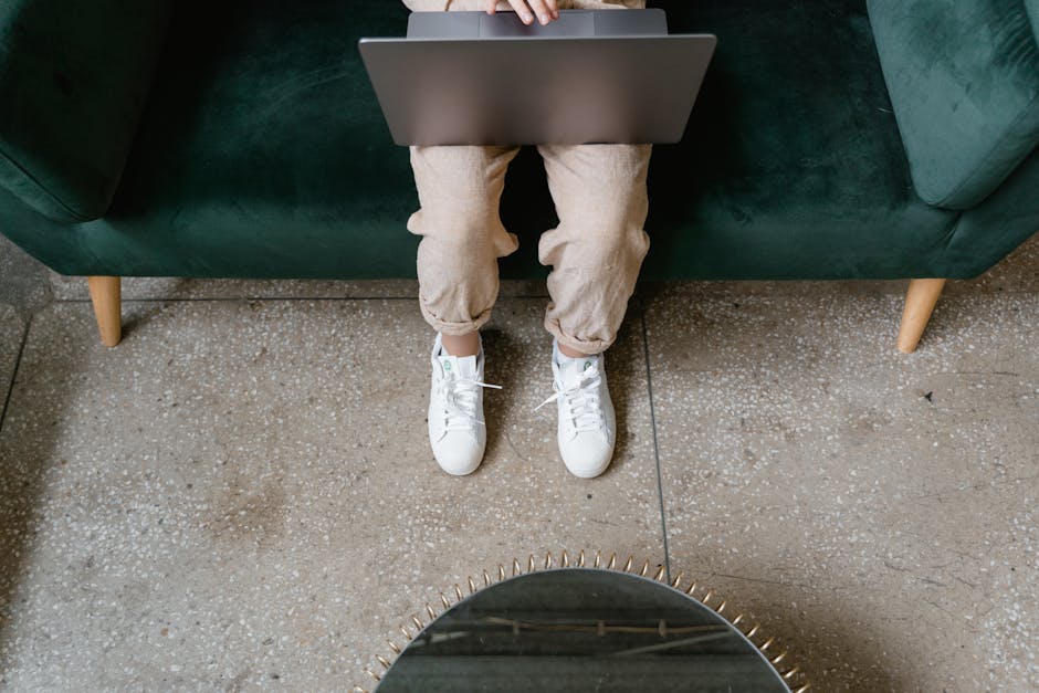 Person sitting on a green sofa using a laptop with white sneakers visible, seen from above.