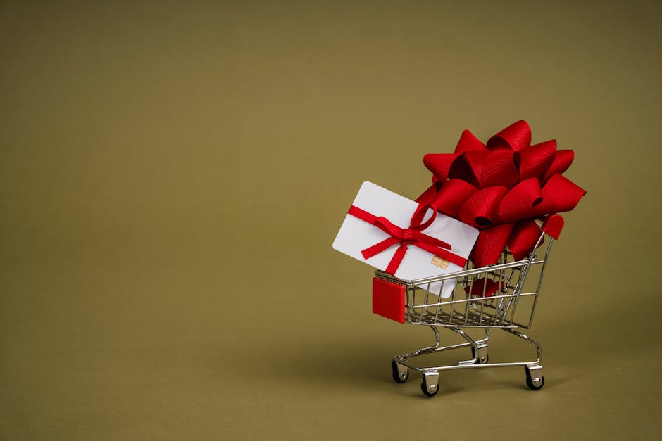 Mini shopping cart containing a gift card adorned with a red bow on a neutral background, ideal for retail concepts.