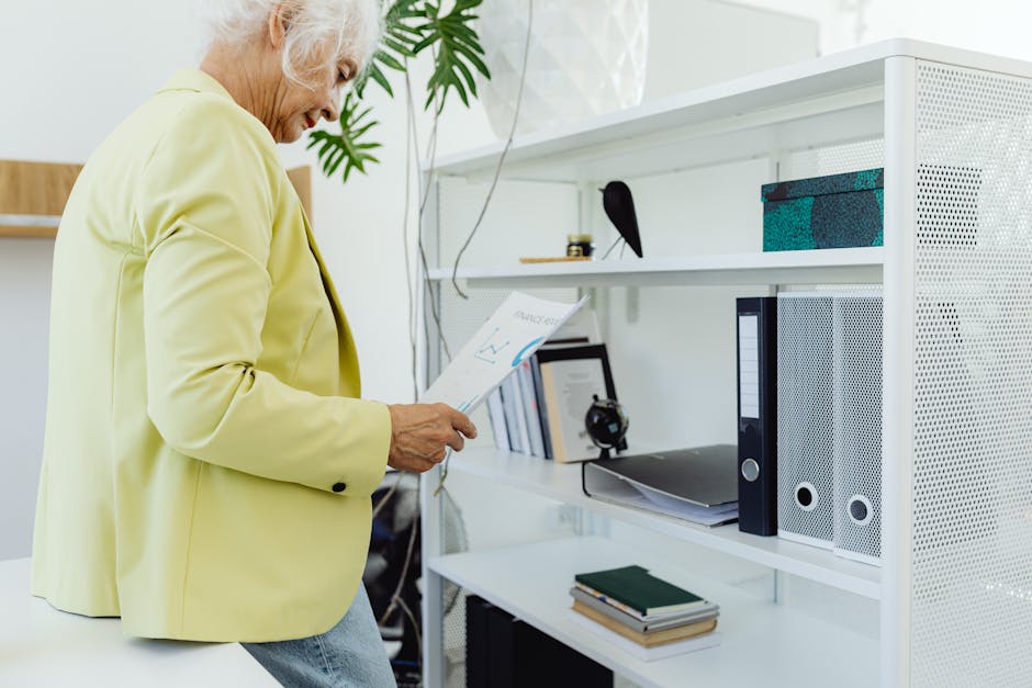 Elderly woman in yellow blazer reviews paperwork while leaning against a white bookcase in an office setting.