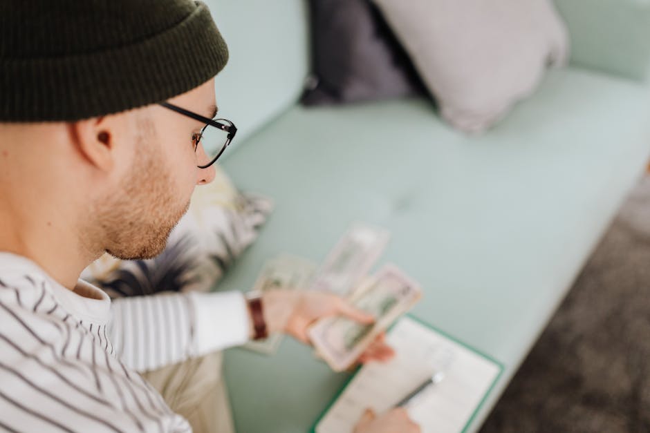 Adult man wearing glasses and beanie counting cash while budgeting indoors.