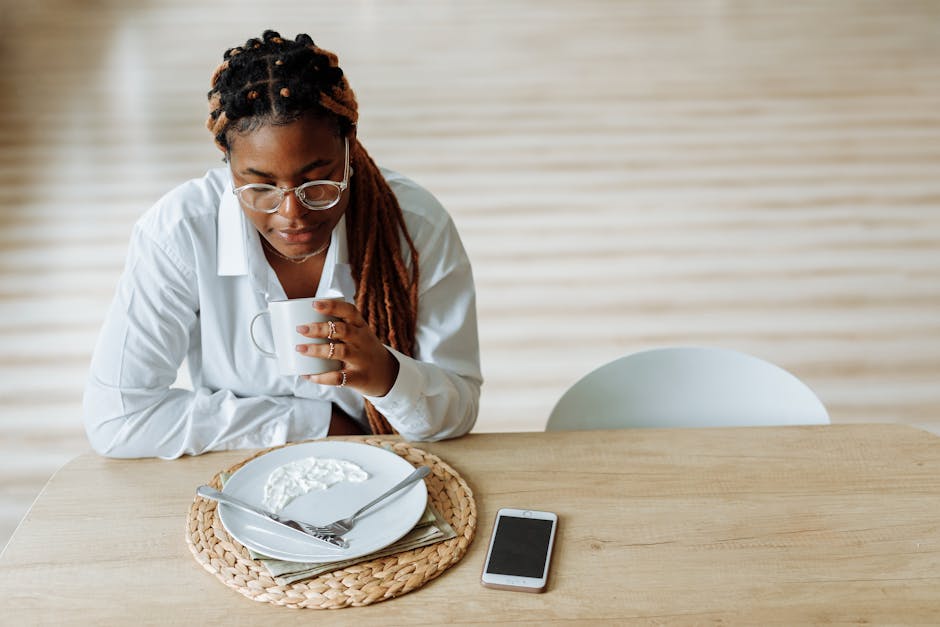 Woman in white shirt enjoying coffee at breakfast with smartphone in view.