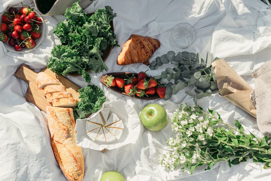 Top view of a vibrant picnic spread featuring fresh fruits, breads, and greens on a white cloth.