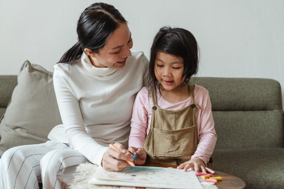 Interested Asian girl in casual outfit leaning on sofa while coloring drawing with tutor using wax crayons in light studio