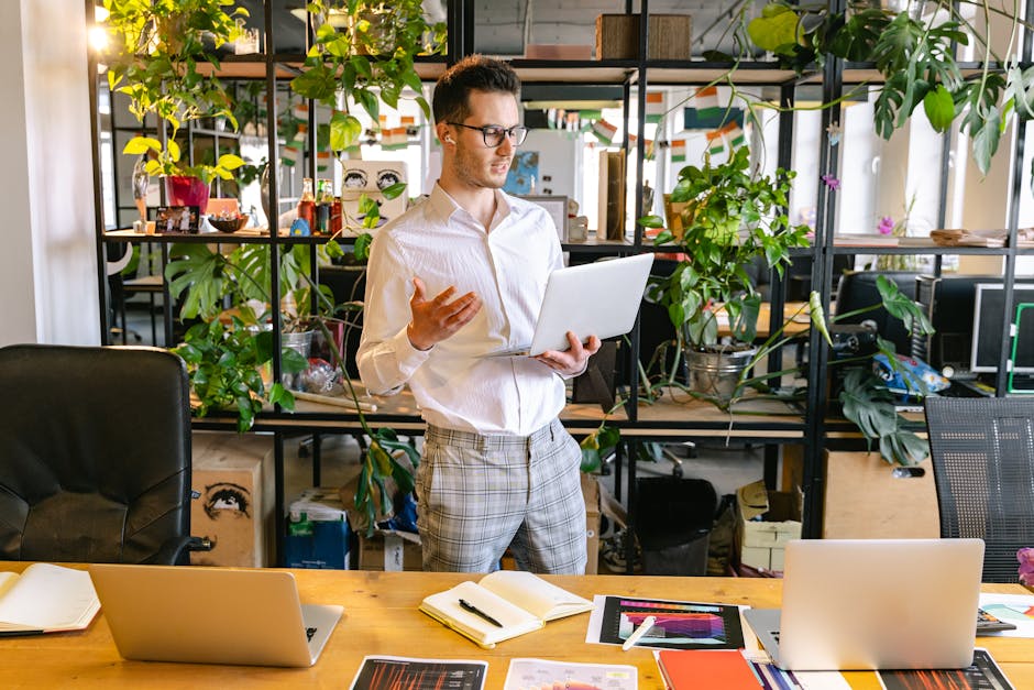 Professional man having a video call in a green indoor office space with multiple laptops and potted plants.