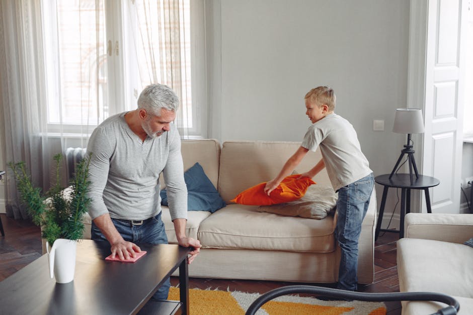 A father and son clean their living room, tidying up cushions and dusting tables.
