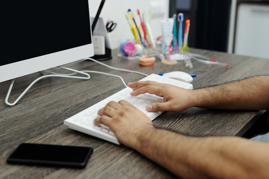 Close-up of hands typing on a white keyboard at a wooden desk in an office setting with accessories.