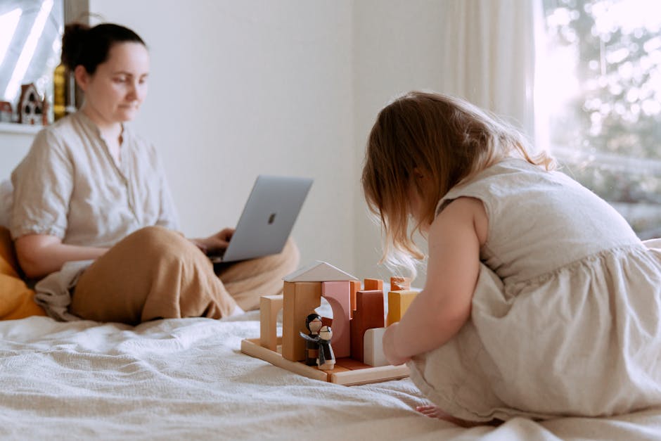 Side view of adorable toddler girl sitting on bed and playing with wooden toys while mother working from home with laptop on knees on sunny morning