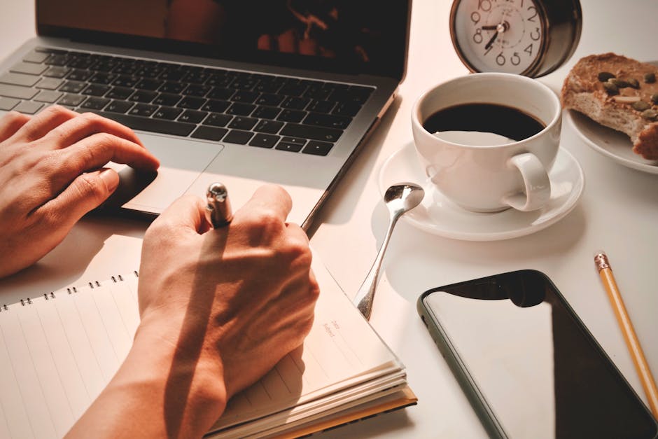 Close-up of hands writing in a notebook with coffee, laptop, smartphone, and alarm clock nearby.