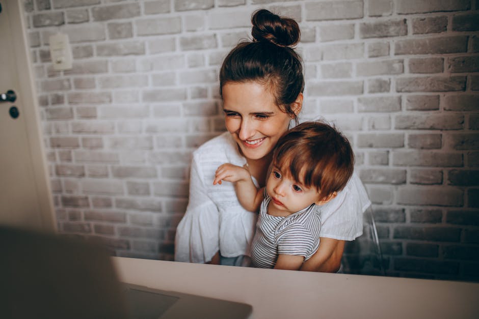 A mother holding her child while smiling at a laptop in a cozy indoor setting.