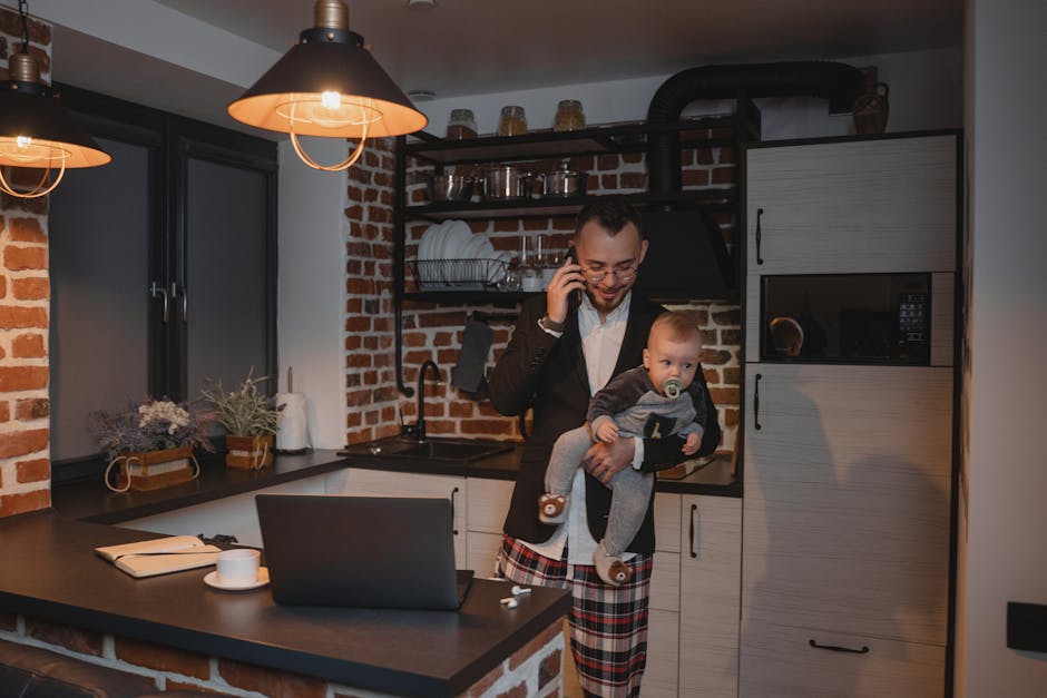 A father multitasks by holding his baby while talking on the phone in a cozy home kitchen.