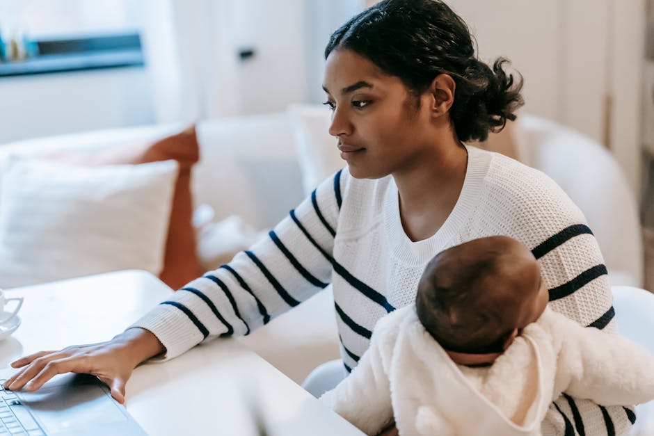 Ethnic baby on laps hugging female in casual outfit with netbook at table while sitting in chair at light apartment near sofa