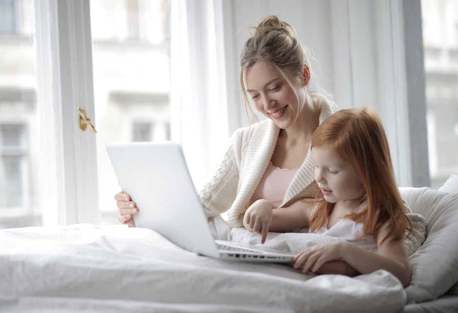 A mother and daughter share a joyful moment in a cozy bedroom, bonding over a laptop.