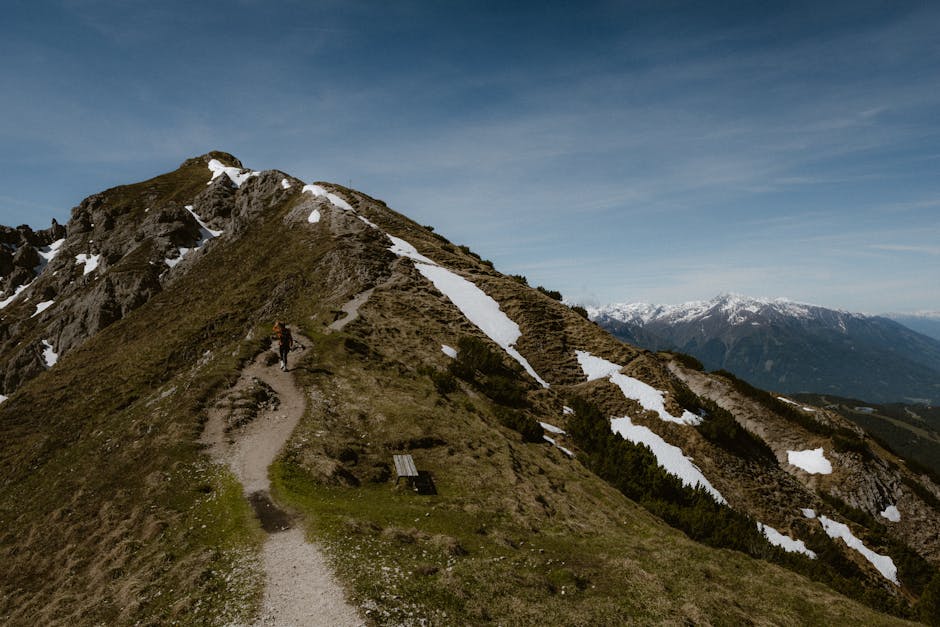 A lone hiker walks along a snowy ridge trail in the Alps on a clear day, capturing adventure and solitude.