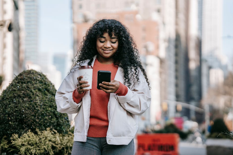 Content African American female in casual outfit standing with coffee to go in hand and using mobile phone on contemporary city street