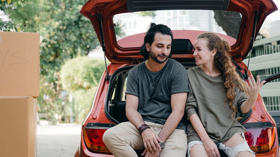 Happy multiracial couple in casual clothes engaged in lively conversation while sitting on edge of luggage boot after unloading carton boxes out of car on sunny day