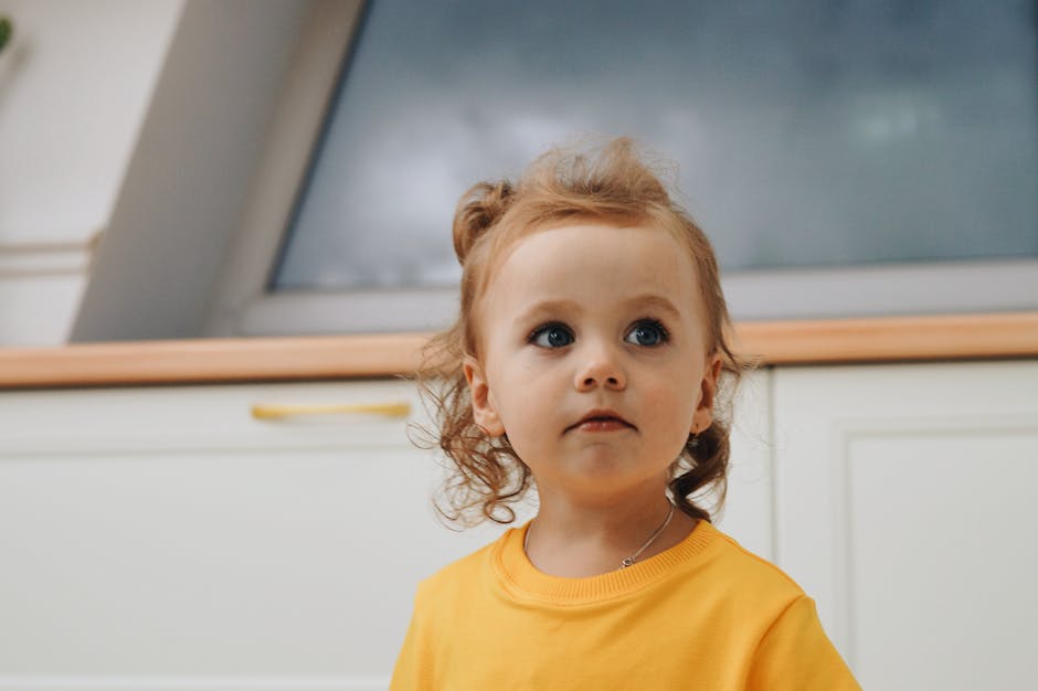 A cute child with curly hair in a yellow shirt looks curiously upwards indoors.