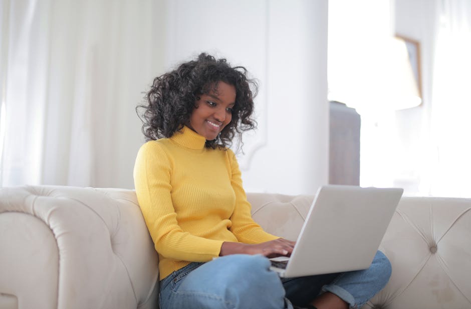 Positive young ethnic lady in casual yellow sweater and jeans sitting on comfortable couch and working on laptop while spending time at home