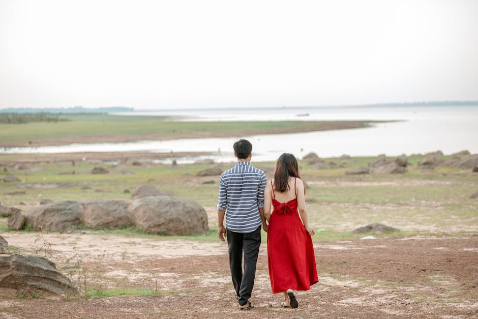 Back view full length couple in stylish outfits holding hands and strolling on sandy rough seashore