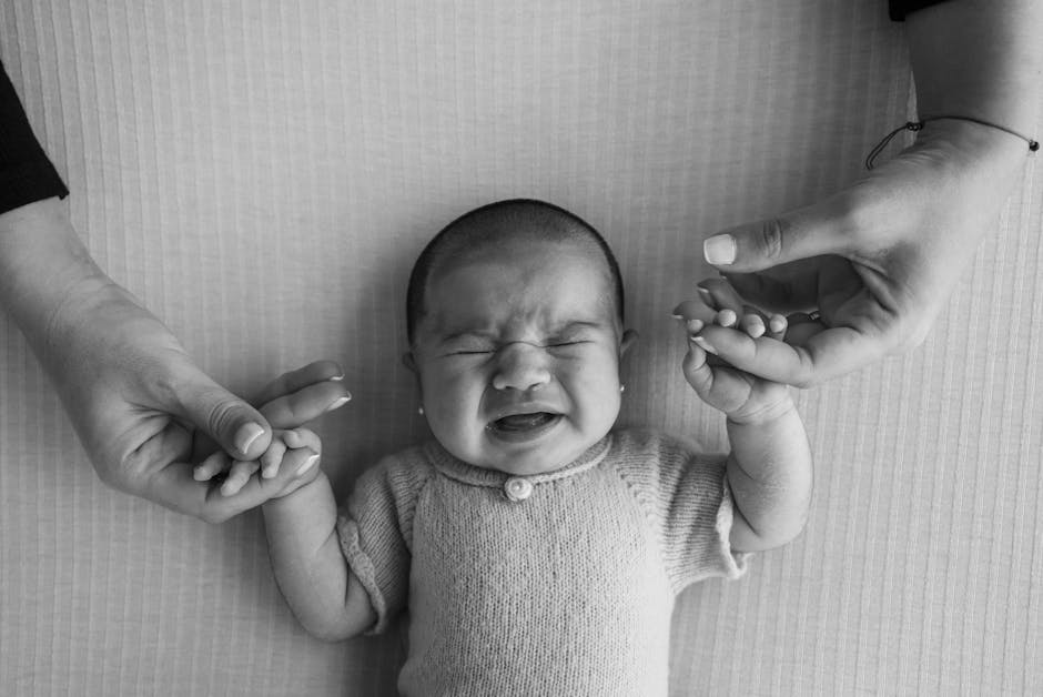 Black and white image of a crying baby being comforted by two adult hands holding theirs.