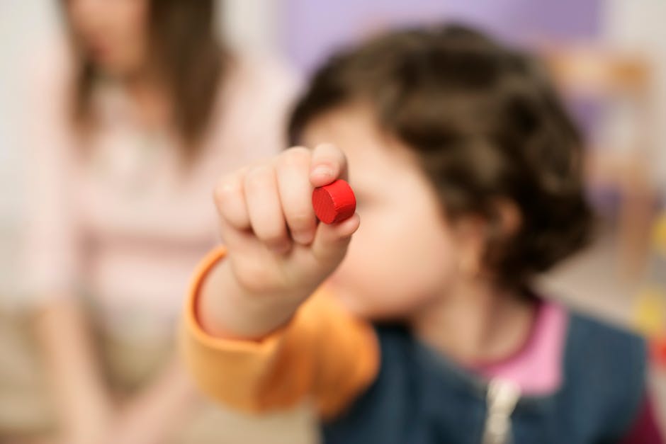Close-up of a child holding a red wooden toy block with a blurred background indoors.