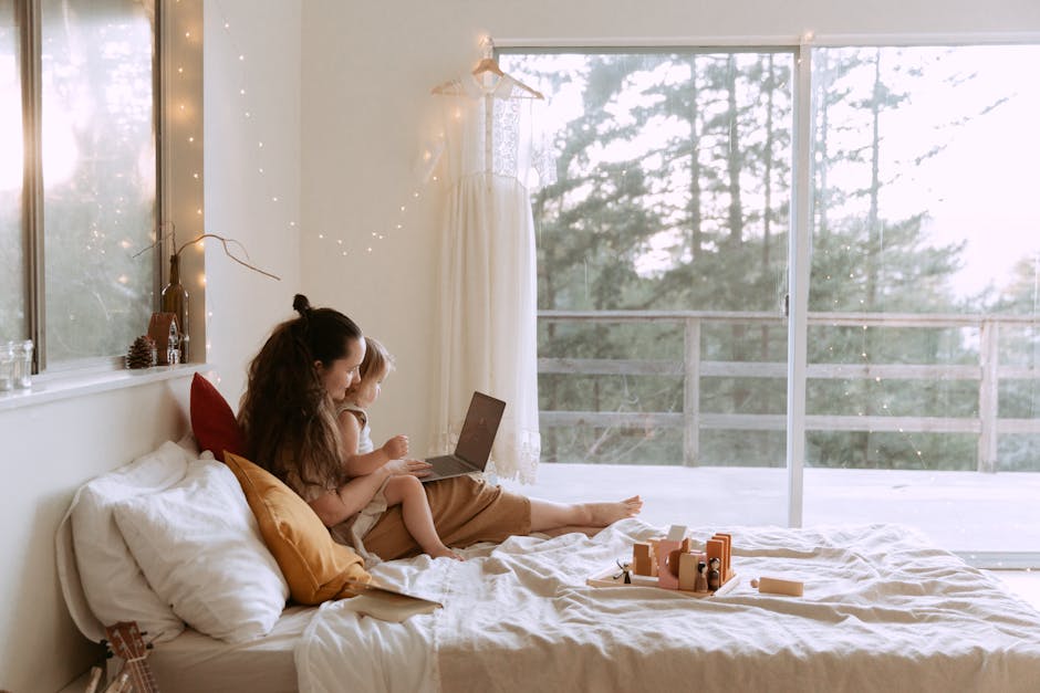 Side view of loving mother and small daughter resting together on bed with laptop and toys