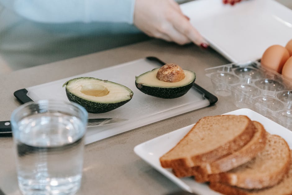 Crop faceless female at table with cut avocados and toasts near water in light kitchen during breakfast breakfast at home