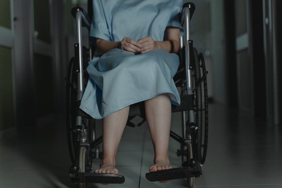 Person sitting barefoot in a wheelchair wearing a hospital gown in a dim hallway.