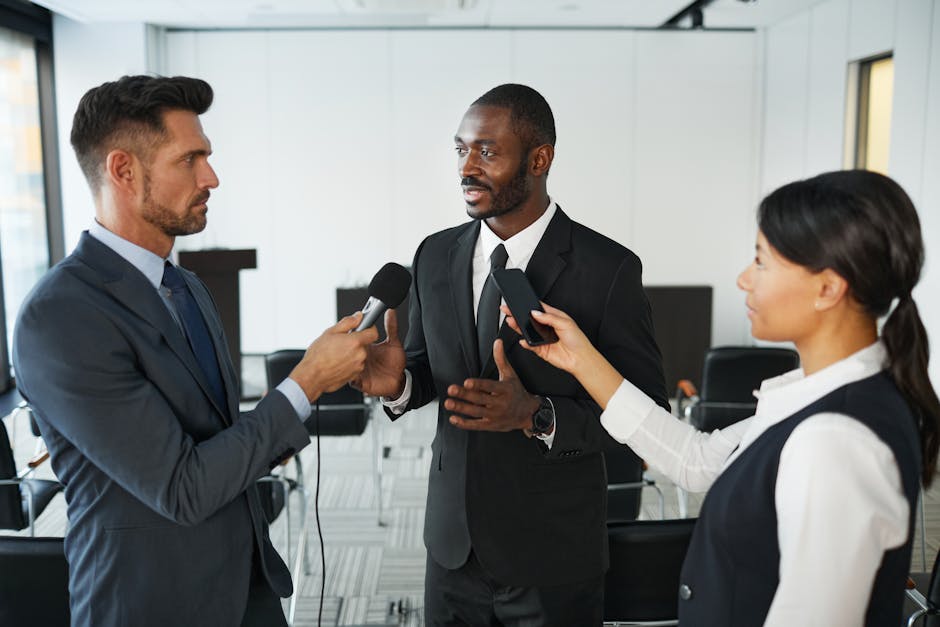 African American businessman in formal attire being interviewed by journalists with microphones in a conference room.