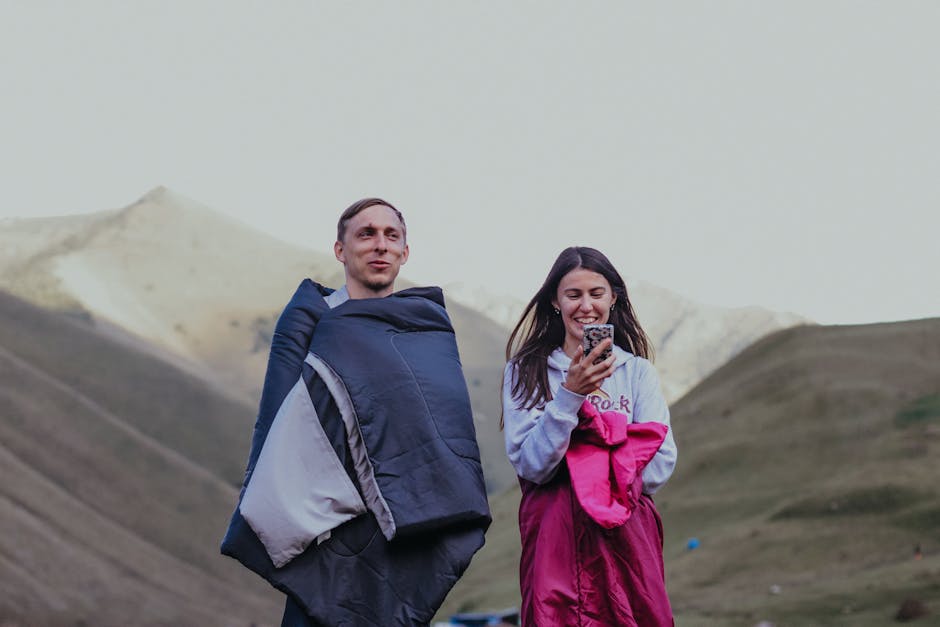 A man and woman wrapped in sleeping bags enjoying nature and outdoor camping in mountainous terrain.