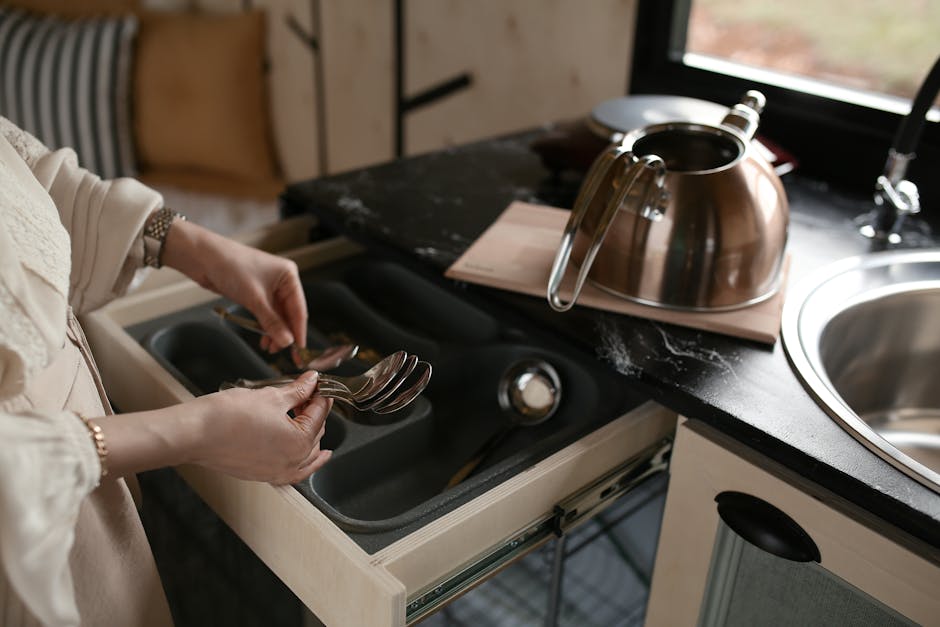 A woman organizing spoons in a kitchen drawer beside a stainless steel kettle.