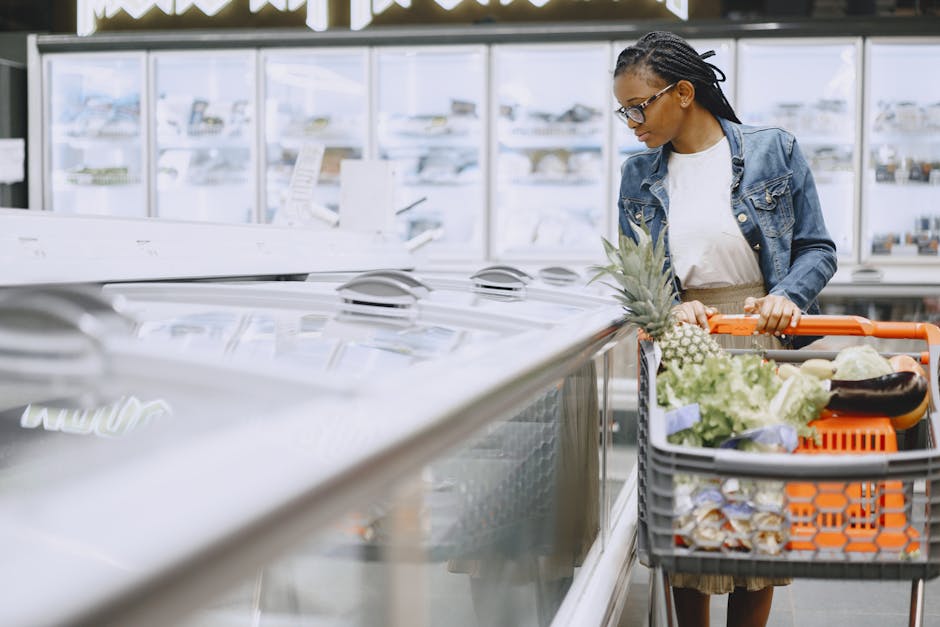 Woman shopping in supermarket with full cart of groceries wearing eyeglasses and denim jacket.