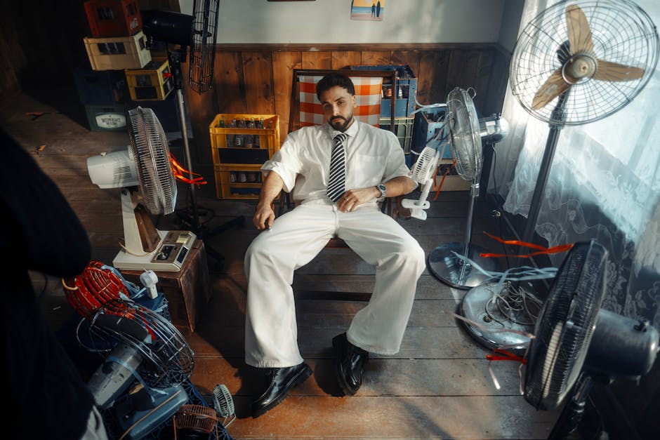 Man in white attire sitting in vintage room with multiple fans and crates. Warm lighting adds classic feel.