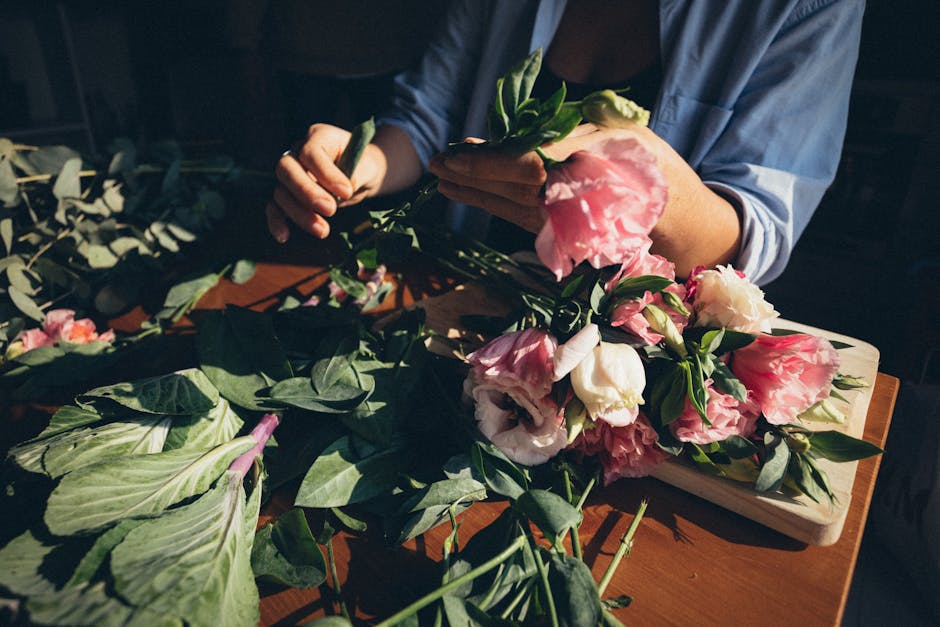 A florist arranging a beautiful bouquet of pink and white flowers using green leaves on a wooden table.