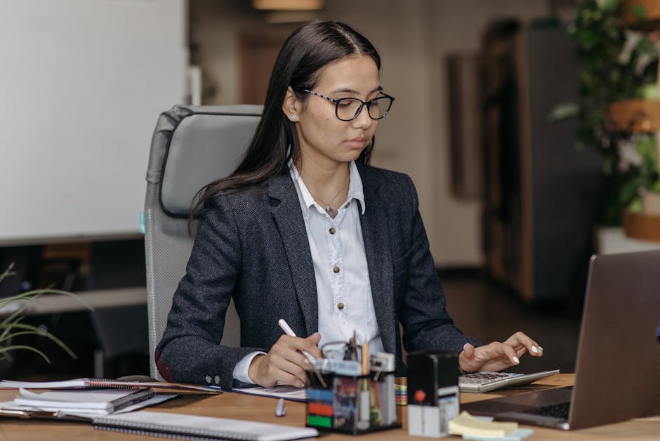 Young woman working with concentration at her laptop in a modern office setting, wearing eyeglasses.