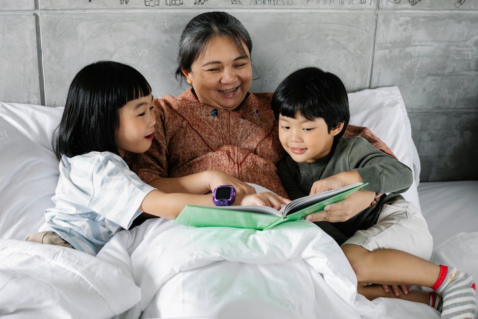 Cheerful grandmother lying under blanket and reading interesting book for Asian children in bedroom
