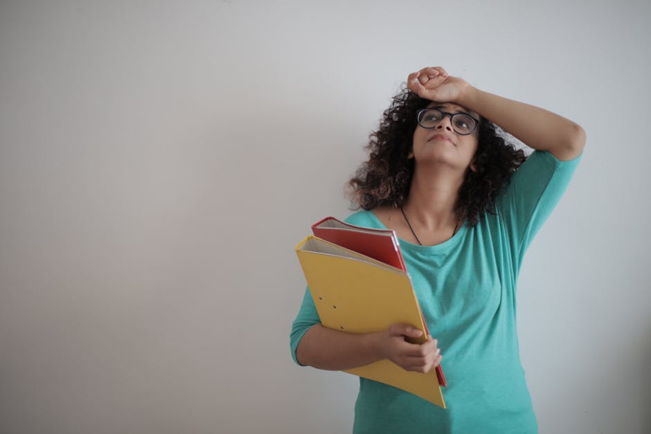 Low angle of unhappy exhausted woman in glasses and casual clothes with folders of papers looking up with sorrow and touching forehead while standing against white wall and having headache due to workload in office
